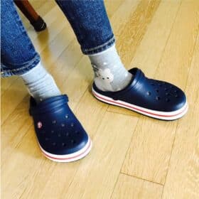 En person i jeans og grå bjørnedesignede sokker står på et trægulv iført Crocs Sandal Børn, med marineblå træsko med hvide og rødstribede såler.